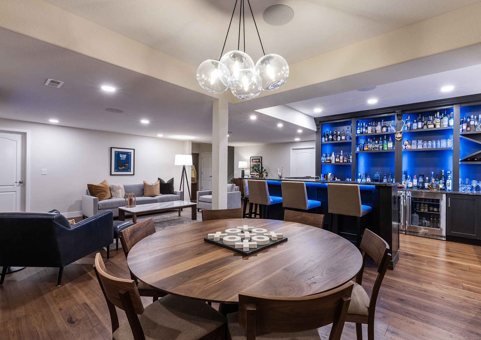 A modern basement lounge featuring a round wooden dining table with chairs, a bar with blue lighting and various bottles, a sofa area with decorative pillows, and a chandelier.