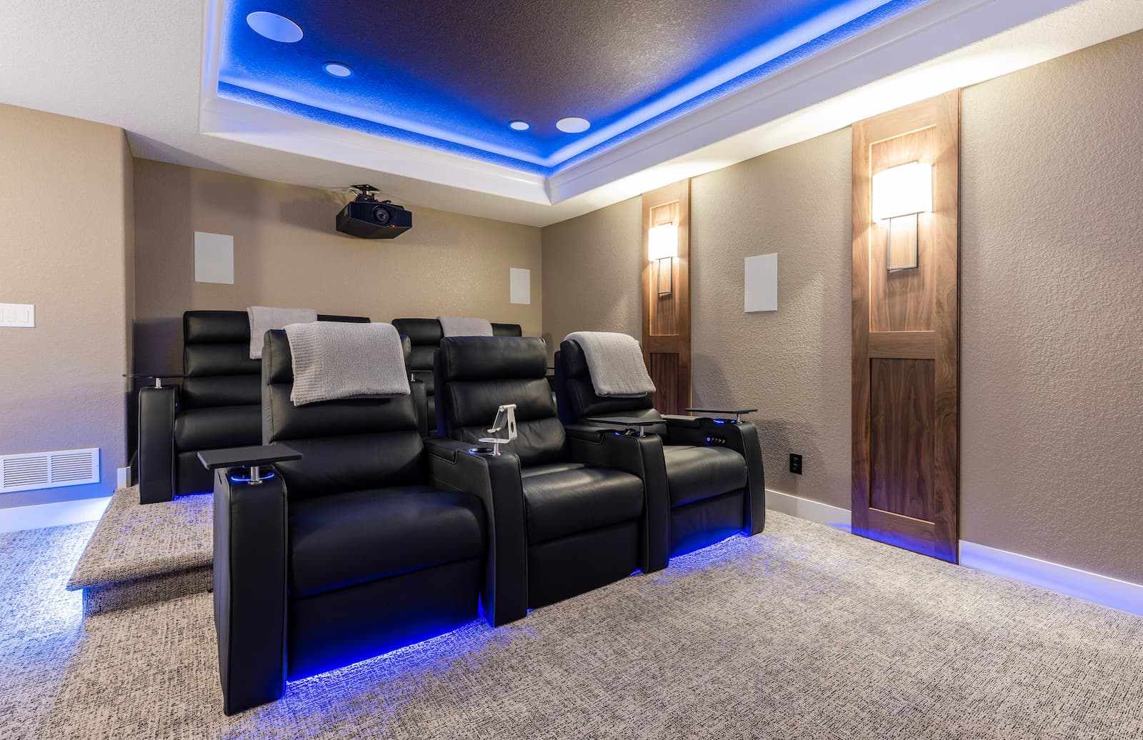 A modern home theater with black reclining chairs, side tables, a projector, and blue ambient lighting.