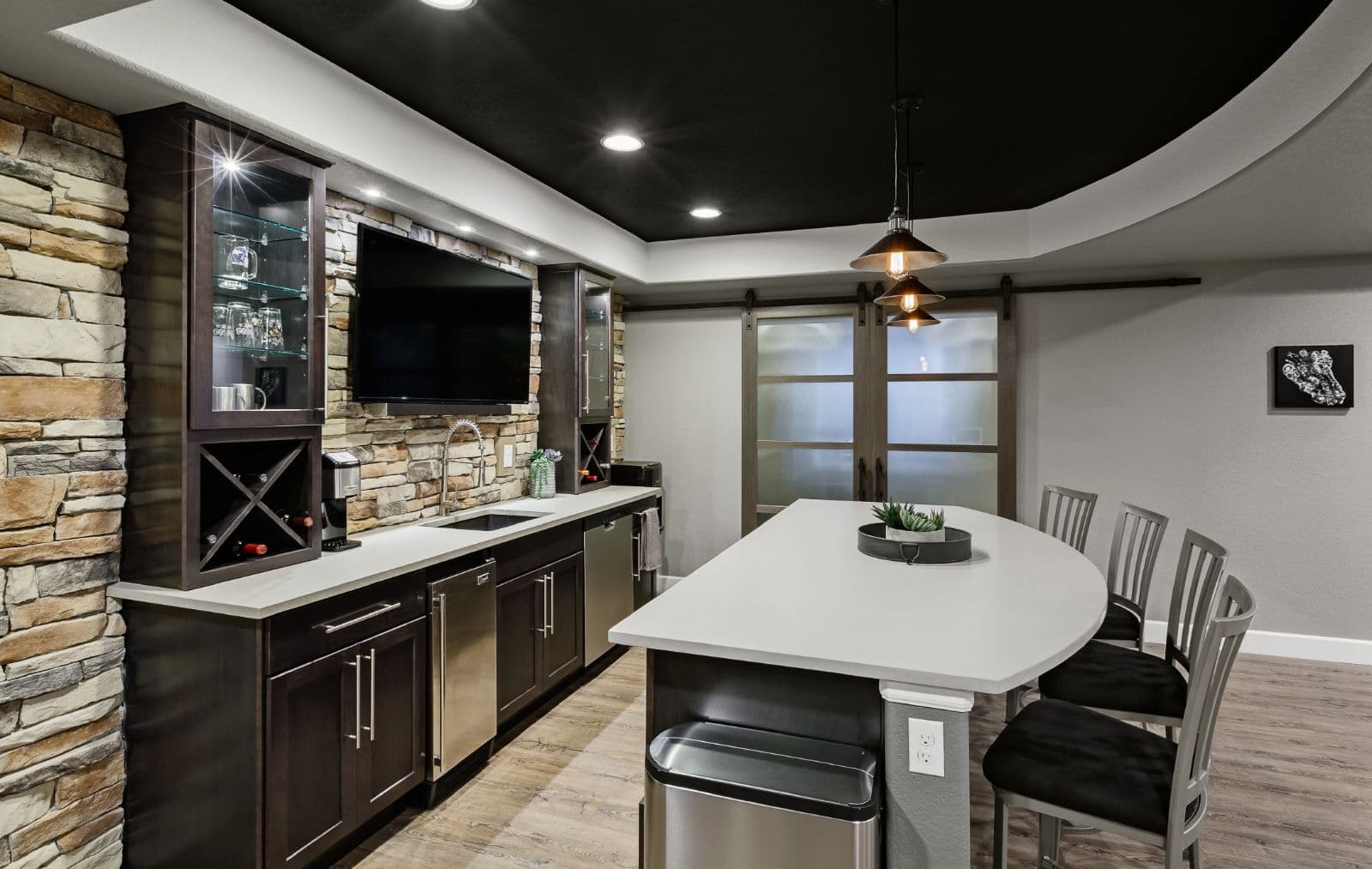 A modern kitchen bar area featuring a stone accent wall, stainless steel appliances, a large countertop, and pendant lighting. There’s a TV mounted on the wall and sliding glass doors in the background.