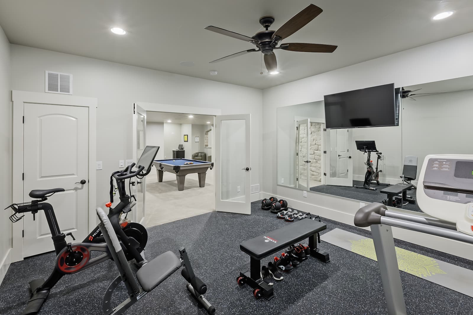 A modern home gym featuring exercise equipment like a stationary bike, weight bench with dumbbells, treadmill, and a mirror. A pool table is visible in an adjoining room.