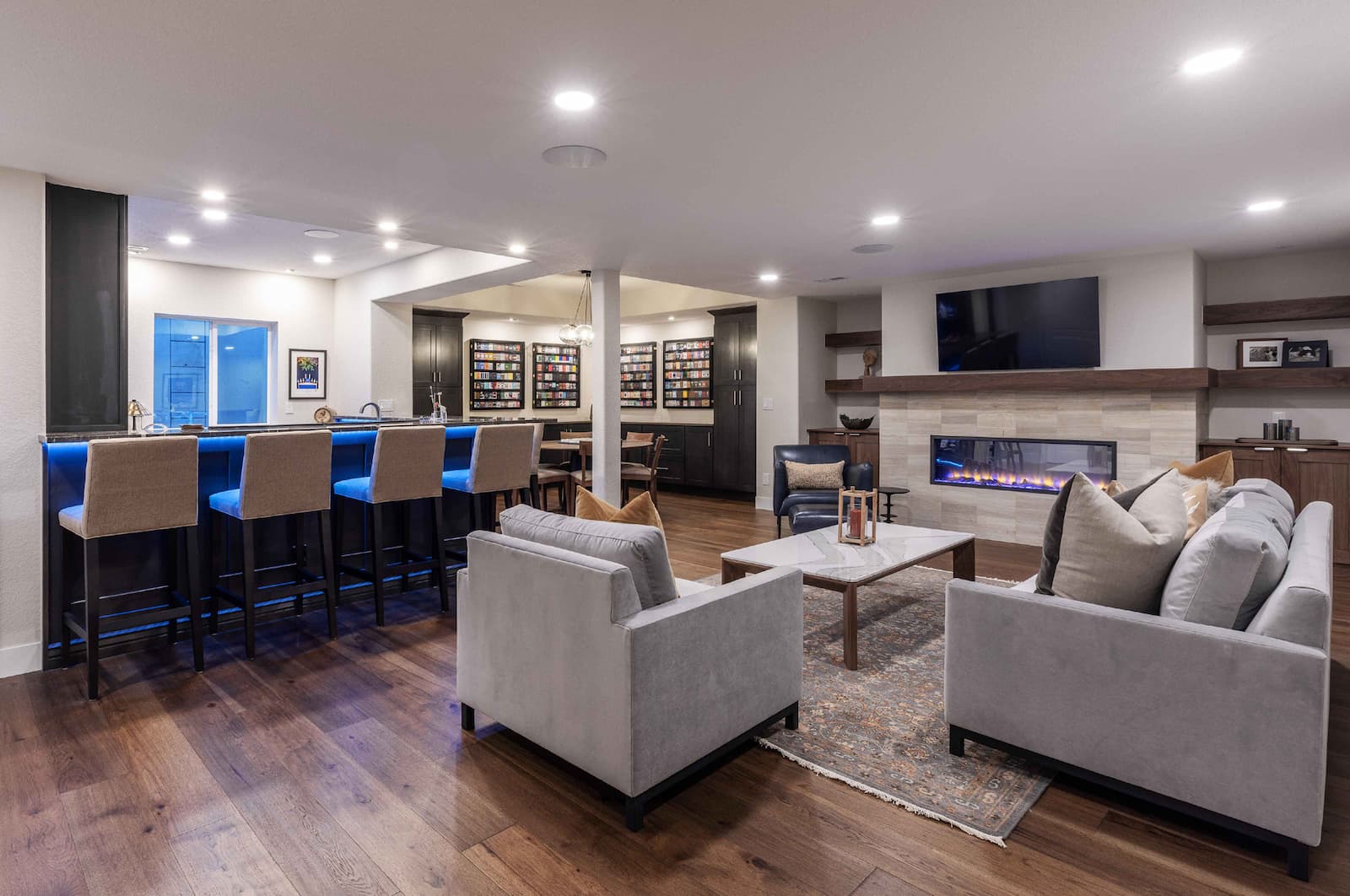A modern living room featuring a cozy seating area with gray sofas, a coffee table, and a decorative rug. There’s a bar counter with high stools, a kitchen area, a fireplace, and shelves with decor. Soft lighting creates a welcoming atmosphere.