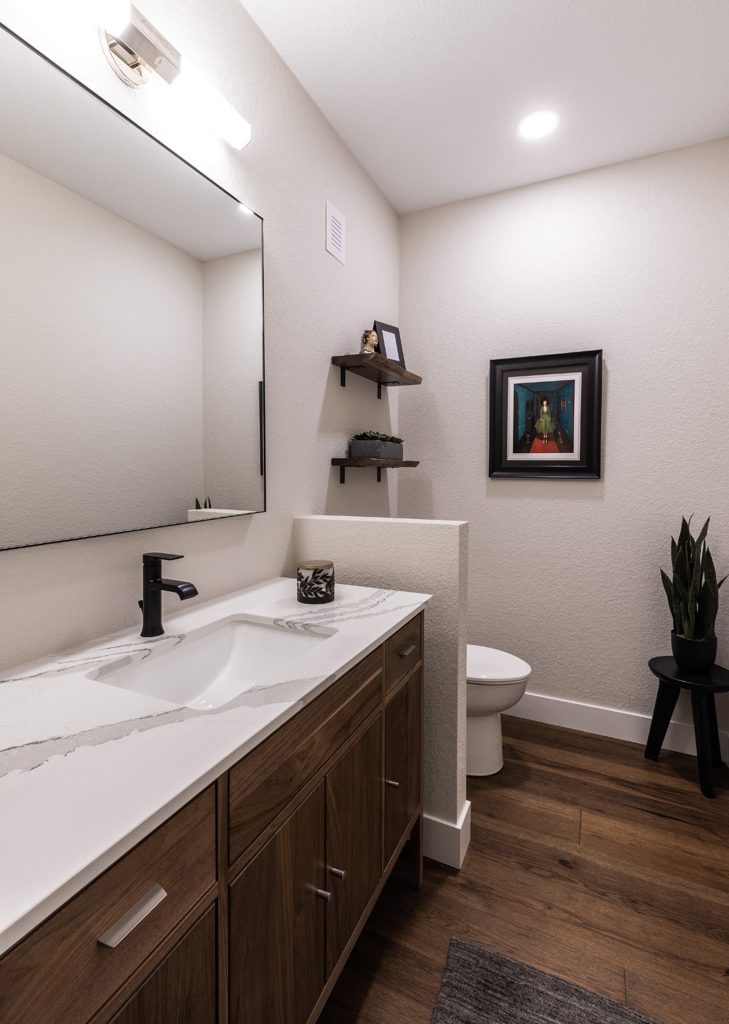 A modern bathroom featuring a white marble sink and countertop, a wooden vanity with drawers, a round mirror, two wall shelves with decor, a framed picture, a toilet, a plant, and wooden flooring.