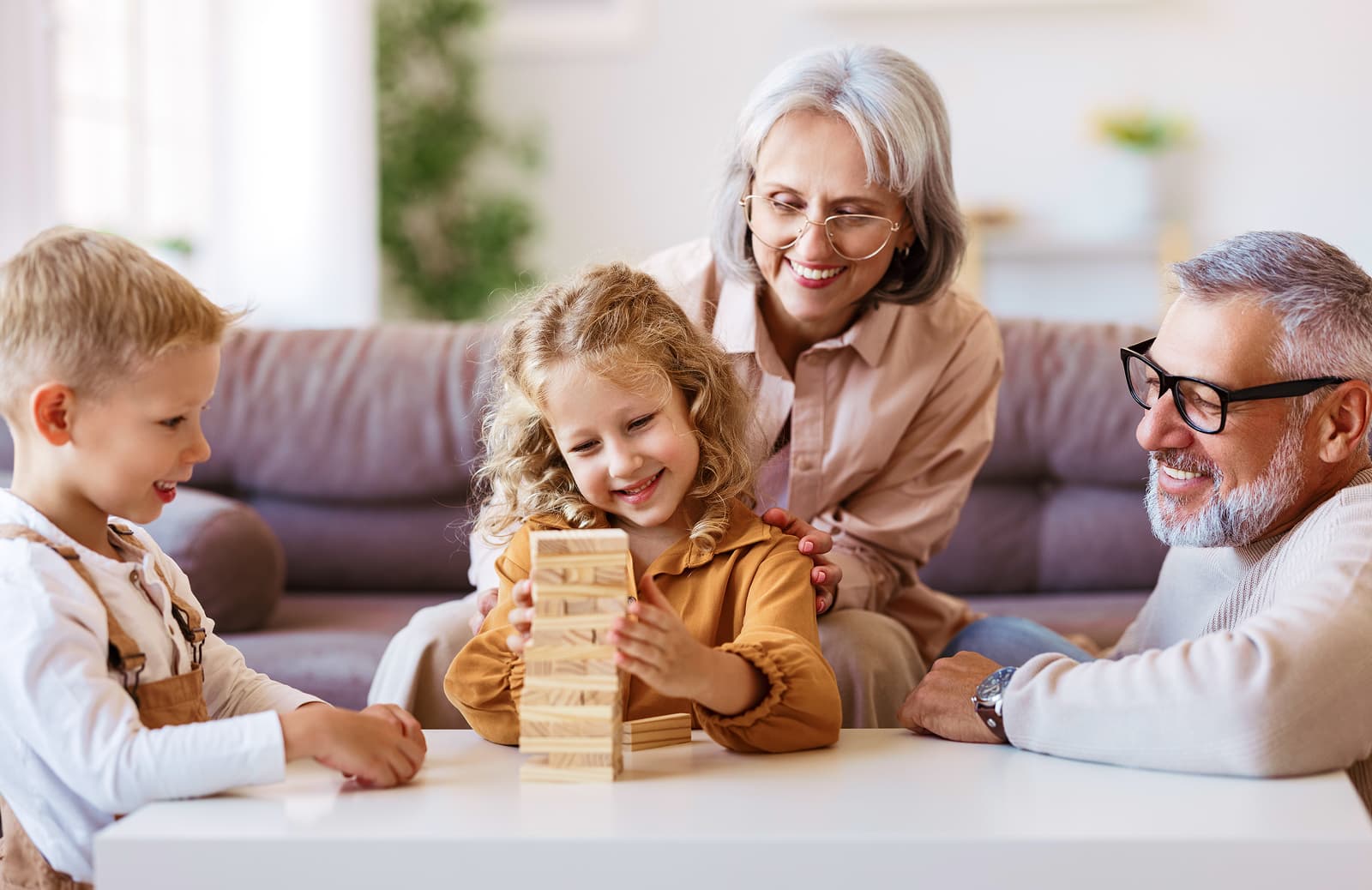 A young girl is happily stacking wooden blocks with the help of her grandmother, while a boy and a grandfather observe and smile, all sitting at a table in a cozy living room.