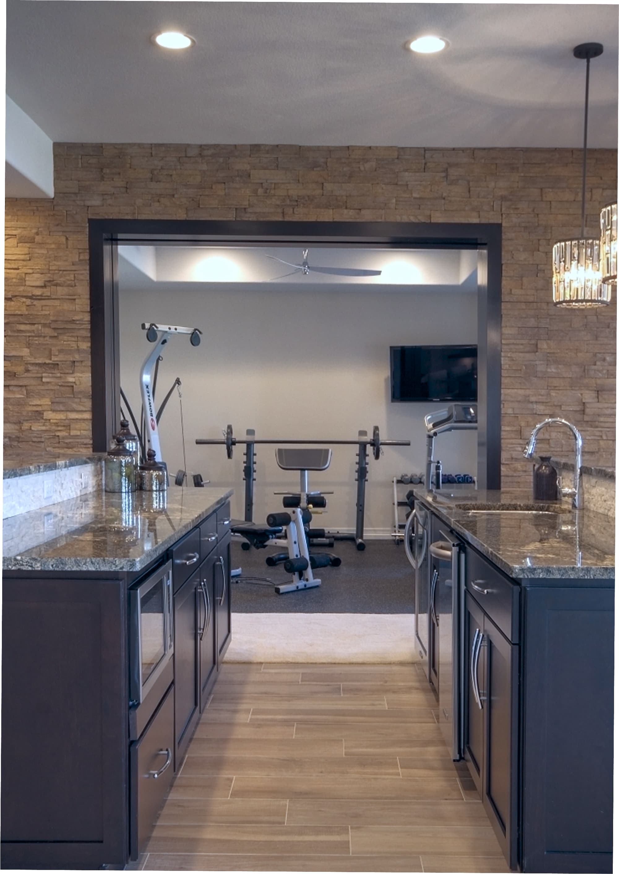 A view of a kitchen with dark cabinetry and granite countertops leading to a home gym featuring exercise equipment and a TV.