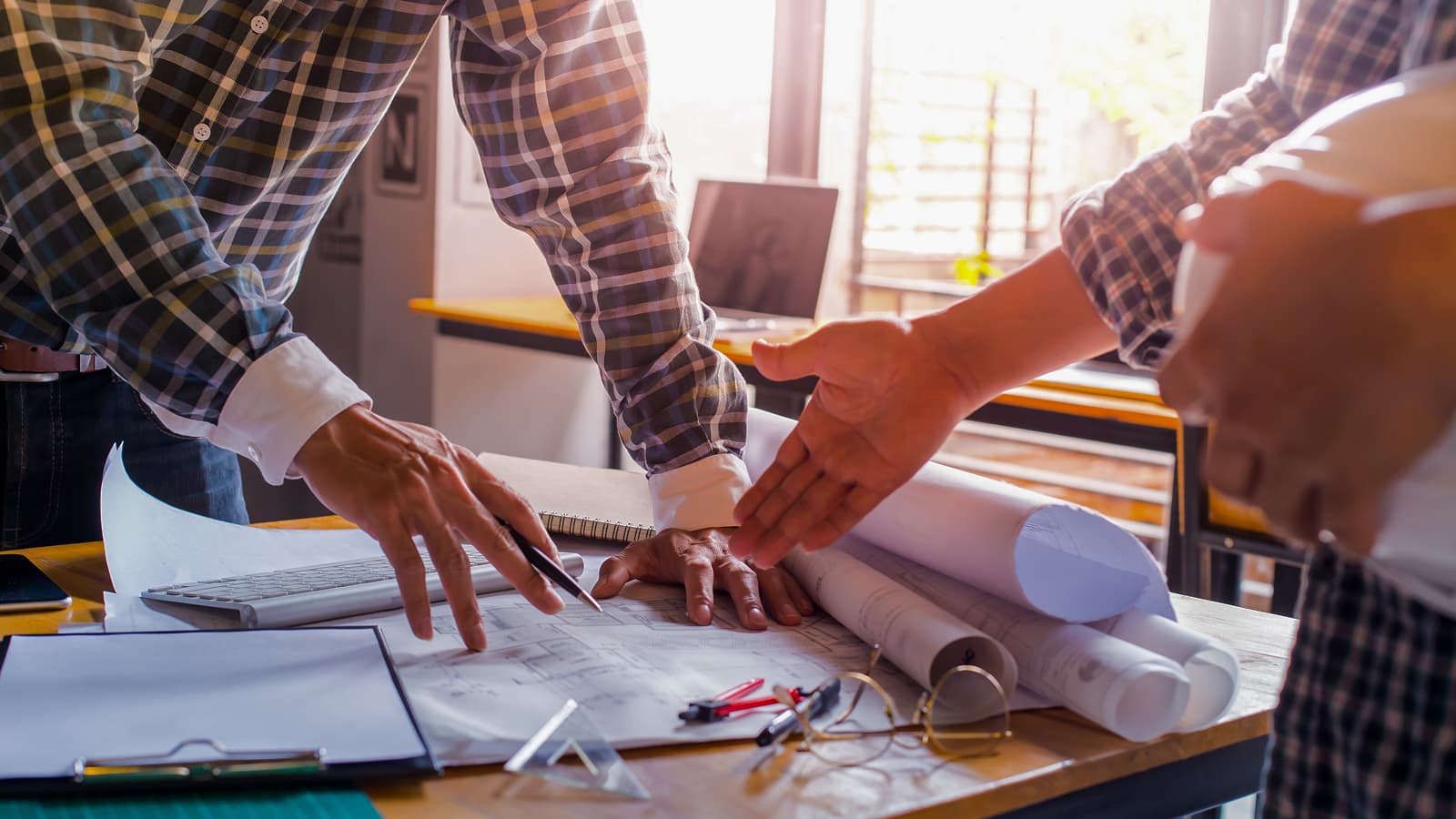 Two people discussing plans at a desk, with hands reaching out and construction blueprints spread out.