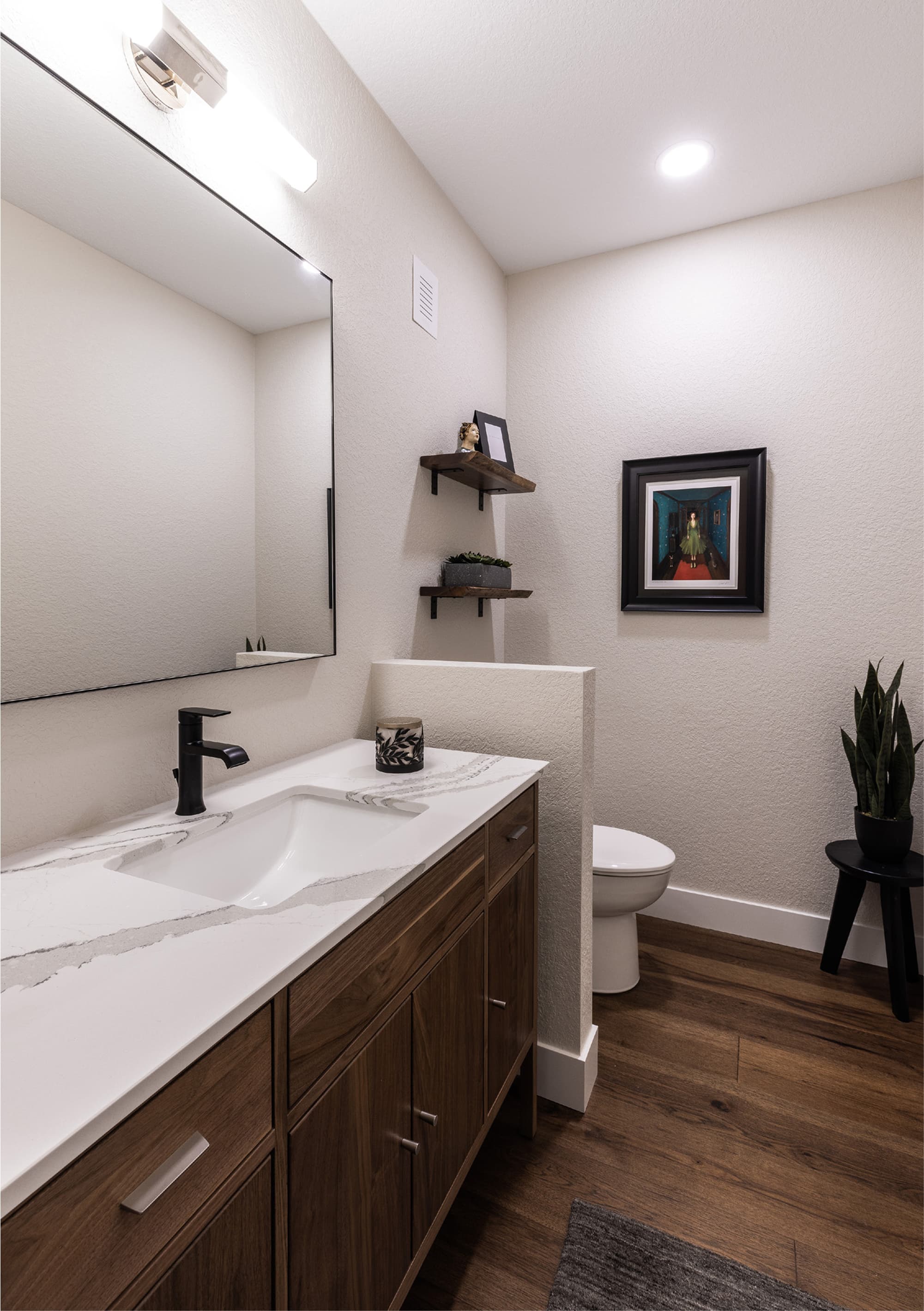 A modern bathroom featuring a white countertop with a sink, a wooden vanity, a large mirror, wall shelves with decorative items, artwork, a toilet, and a plant on a small black table.