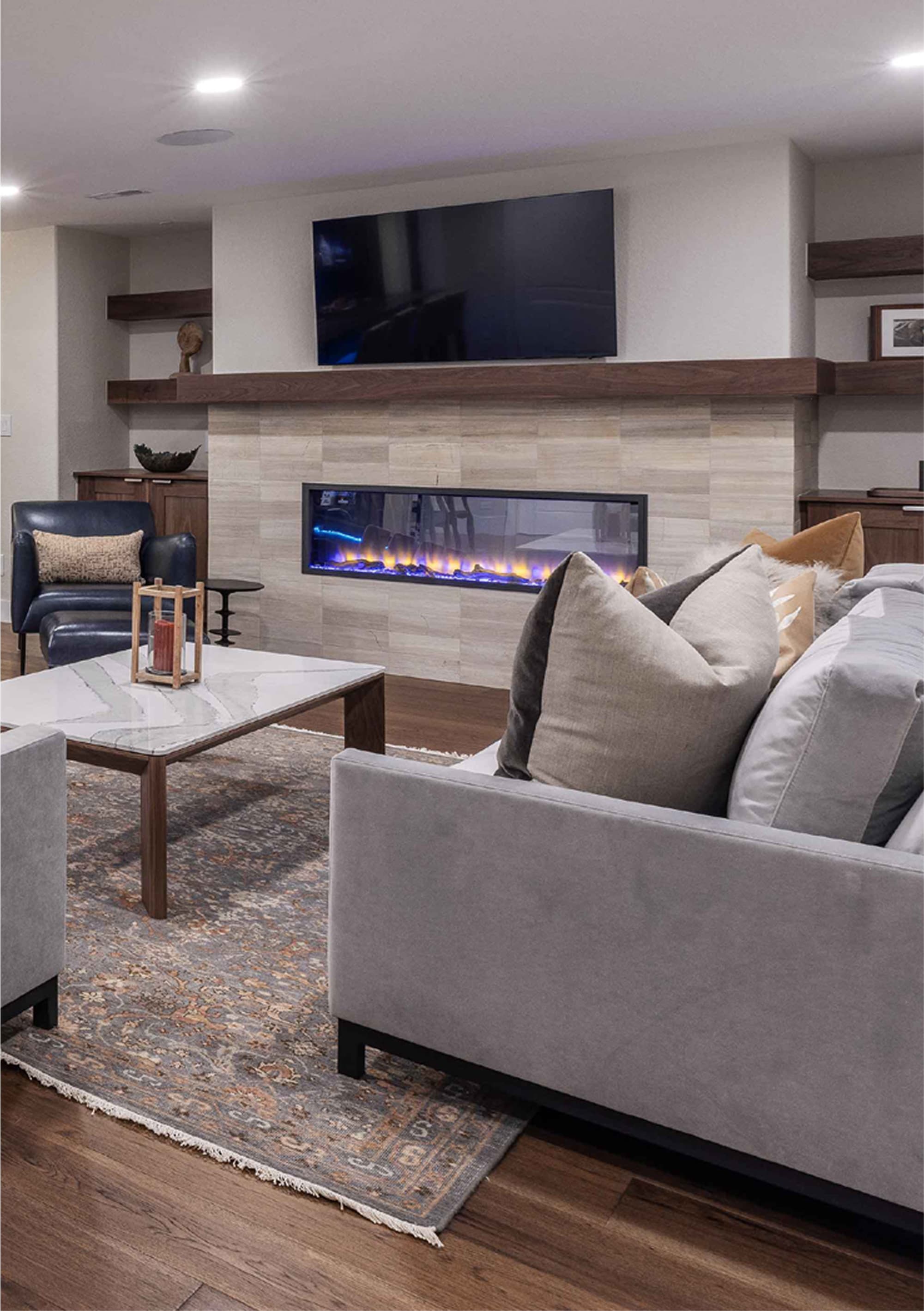 A modern living room with a bar area featuring stools, a coffee table, and a fireplace. The decor includes a wall-mounted TV and shelves with decorative items.