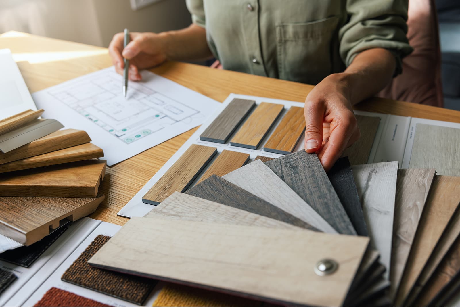 A person selecting material samples, including wood and fabric swatches, while reviewing a floor plan on a wooden table.