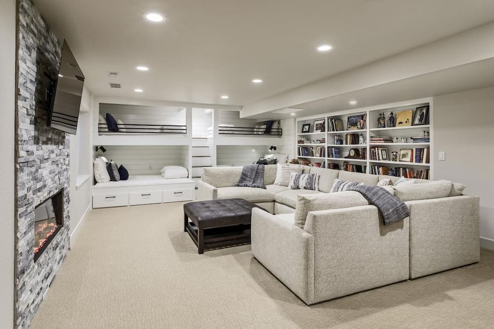 A modern basement living area with a sectional sofa, a dark coffee table, a stone fireplace, and built-in shelves filled with books and decorative items. There are two bunk beds in a nook and soft lighting throughout.