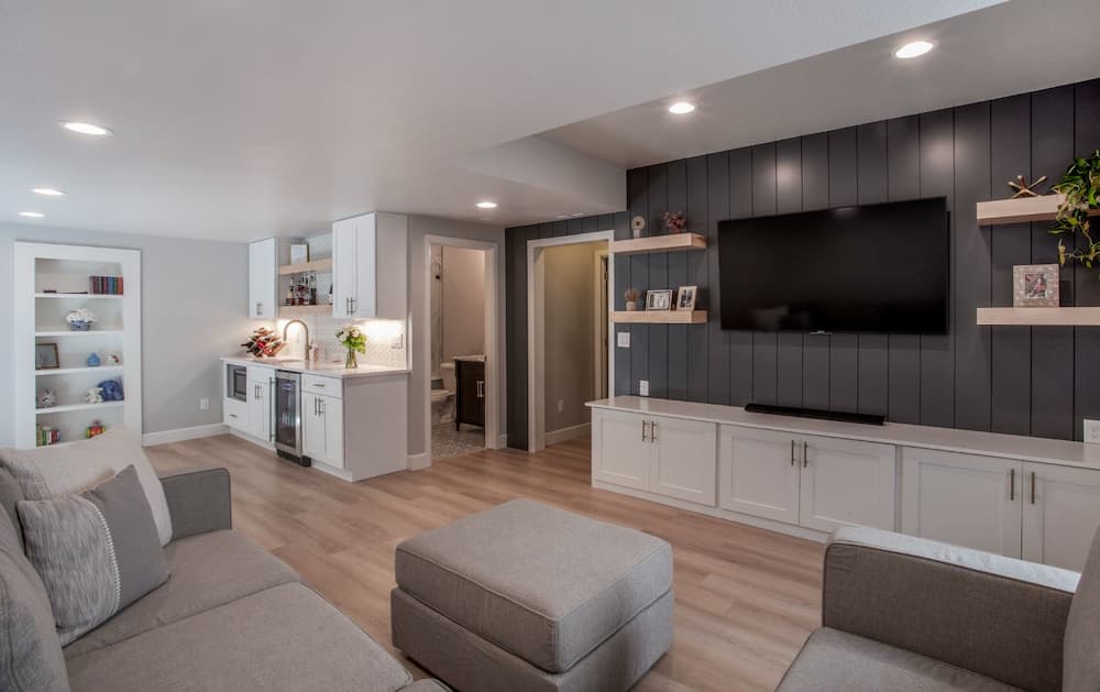 A modern living room featuring a couch, an ottoman, a wall-mounted TV, and a kitchenette with white cabinets and shelves.