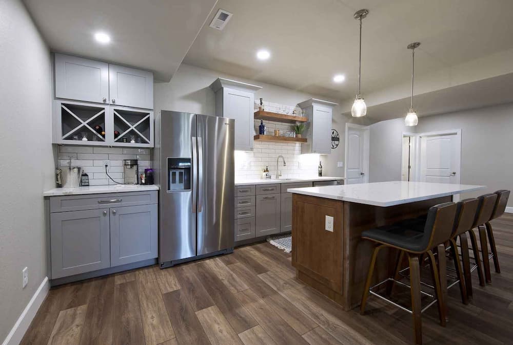 A modern kitchen featuring gray cabinets, a stainless steel refrigerator, a white countertop, and a large kitchen island with seating. The space has pendant lighting and wood flooring.