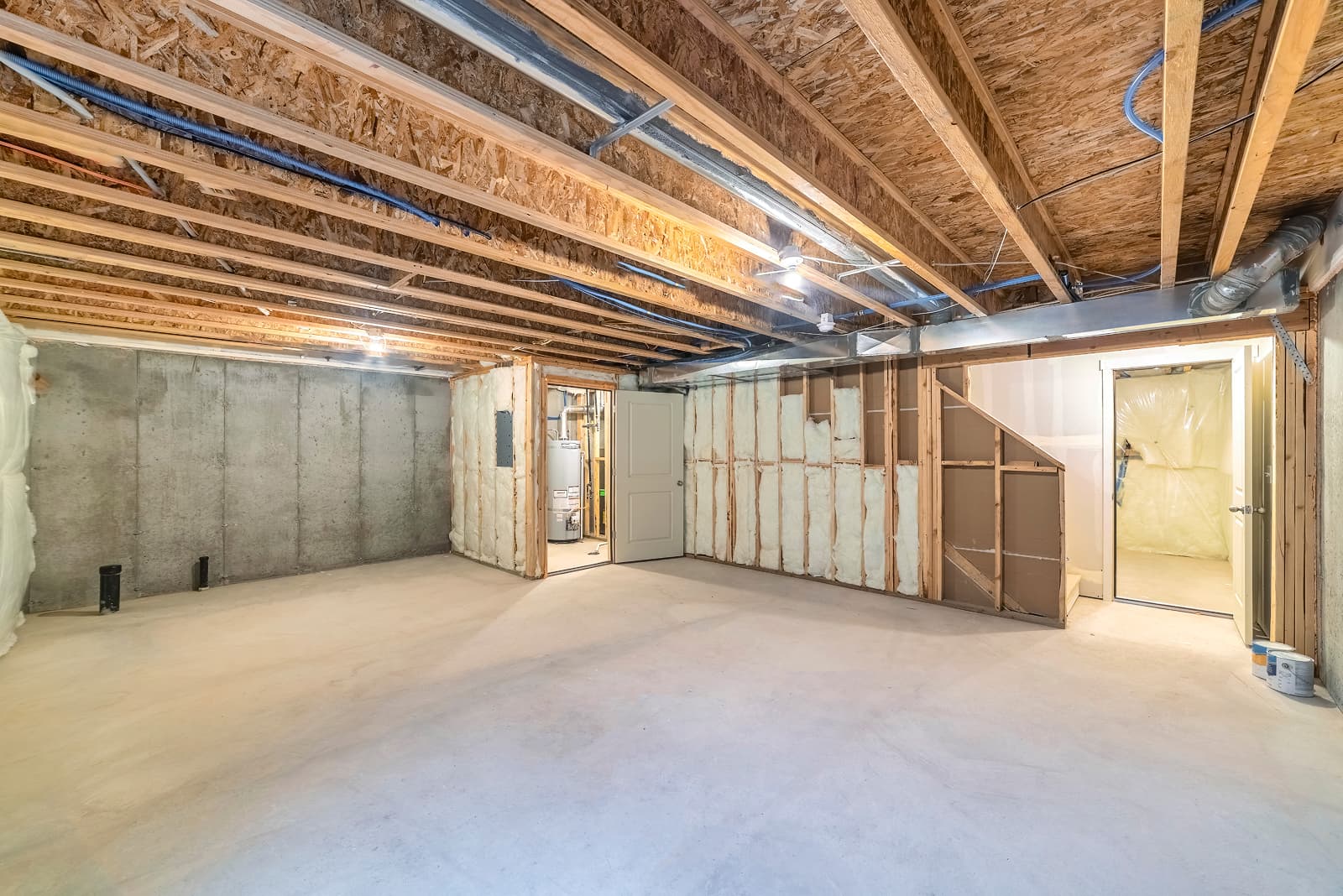 A spacious unfinished basement with exposed wooden beams and insulation on the walls, featuring a door leading to a small room and a concrete floor.